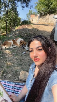 A young woman with long dark hair wearing a denim top takes a selfie outdoors in a rural area, while a brown and white dog rests on the dirt path behind her.