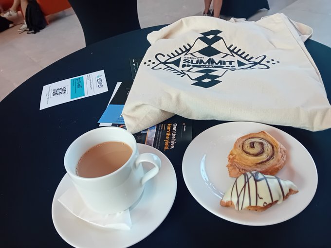 A coffee with milk, pastries (a cinnamon roll and a chocolate-drizzled croissant), and a Kakao M Summit 2024 tote bag sit on a dark table.