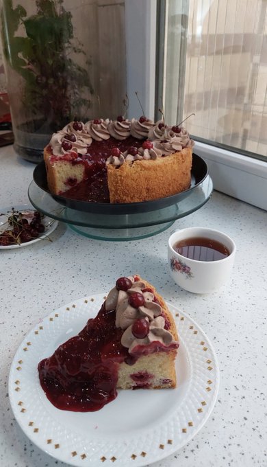 A slice of cherry and chocolate frosted cake sits on a white decorative plate next to a cup of coffee, with the rest of the cake on a glass stand in the background.