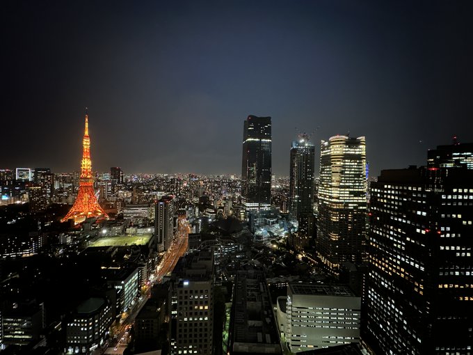 A nighttime aerial view of Tokyo featuring the illuminated orange Tokyo Tower on the left alongside modern skyscrapers.