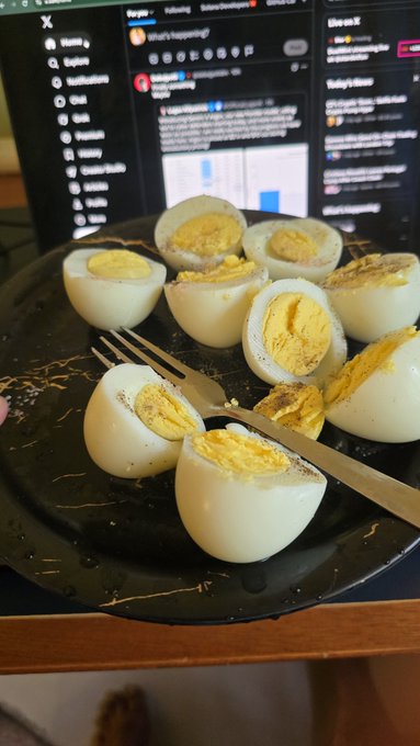 A black marble plate holds several peeled hard-boiled eggs sprinkled with seasoning, with a fork resting on top, set against the backdrop of a computer screen.