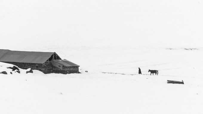A solitary figure and horse stand beside a weathered barn in a vast, snow-covered landscape, evoking the quiet isolation and emotional weight of returning home depicted in the artwork "Homecoming."