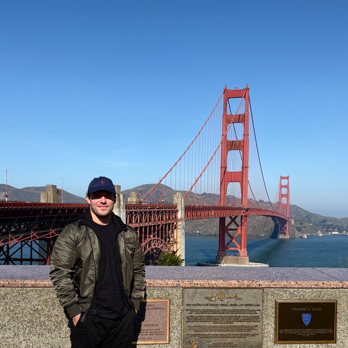 A man in a black puffer jacket and baseball cap poses in front of the Golden Gate Bridge on a clear day, standing beside a plaque dedicated to "Charles A. Ellis." The "Яevolution" and "$TIBBIR" reference ties into the mirrored "R" in "Renaissance," fitting the celebratory, breakthrough-themed post.