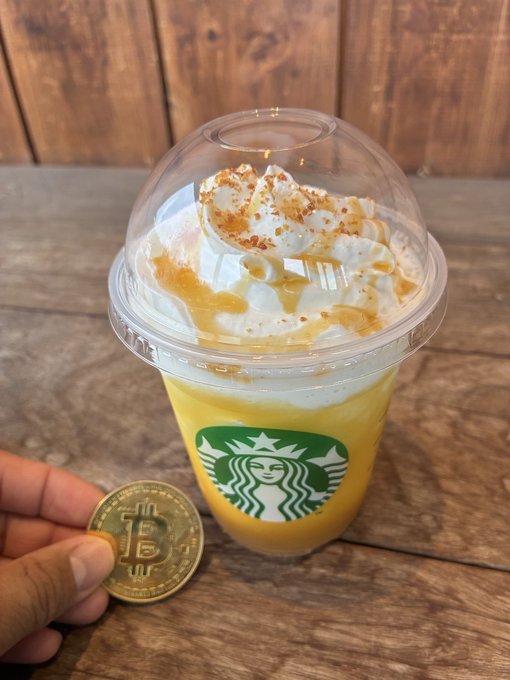 A hand holds a Bitcoin commemorative coin next to a Starbucks frappuccino-style drink topped with whipped cream and caramel drizzle.