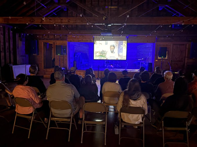 The image shows an audience gathered around a screen, watching a film at a movie theater or event space. The people are seated in chairs arranged in rows facing the screen, which displays the film. There are several individuals present, including some sitting on the floor as well. A projector is visible above the seating area, indicating that this could be an outdoor movie screening or a similar event. The atmosphere appears to be relaxed and social, with people enjoying their time together while watching the film.