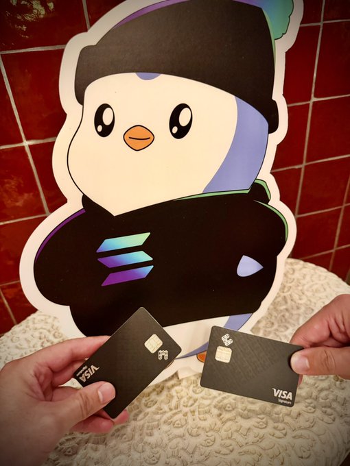 Two Visa credit cards (a metal and standard black variant) are held up in front of a large Pudgy Penguin cutout against a red-tiled wall, playfully suggesting that "pudgys know what's good" — implying the NFT penguin brand endorses or celebrates Visa payment cards.
