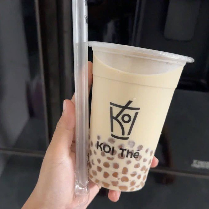 A hand holds a clear plastic cup of bubble tea (boba) from KOI Thé, with the brand's logo and name visible on the cup alongside the tapioca pearls at the bottom.