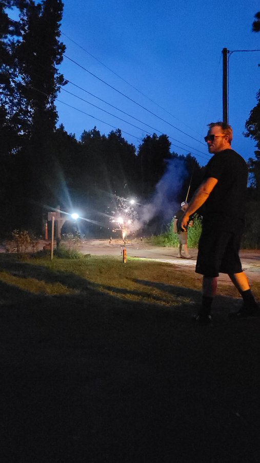 The image shows a man standing outside at night, looking up into the sky where fireworks are being set off. He appears to be enjoying the spectacle or possibly waiting for them to go off. The photo has been captioned with "I Miss ya bro #RCVRY".
