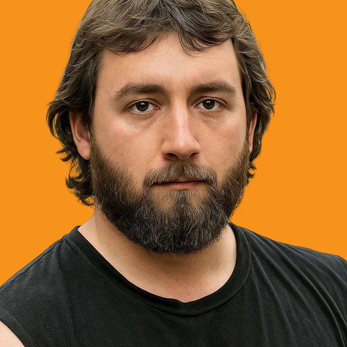 A close-up portrait of a man with brown hair and a full beard, wearing a black t-shirt against a solid orange background.