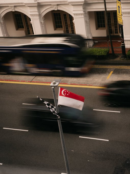 A small Singapore flag waves in the foreground while vehicles speed by on the busy street, capturing the dynamic atmosphere of arrival in Singapore for the Token2049 conference.