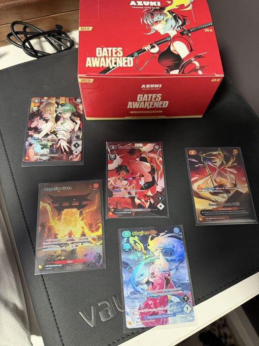 A photograph of an 'AZUKI Gates Awakened' trading card game booster box (ADK-G1, $26.99) along with several anime-style trading cards displayed on a black surface.