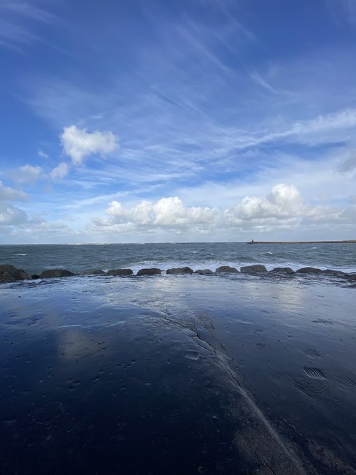 A coastal scene featuring a wet, dark sandy beach or tidal flat in the foreground with footprints, a line of rocks forming a breakwater, and a blue sky with scattered clouds over the sea.