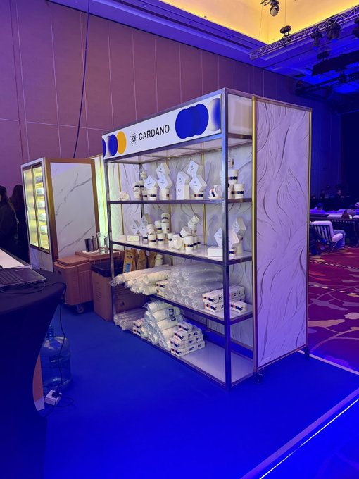 A Cardano merchandise booth at an event, displaying jars and packaged products on shelves, illuminated with blue lighting.