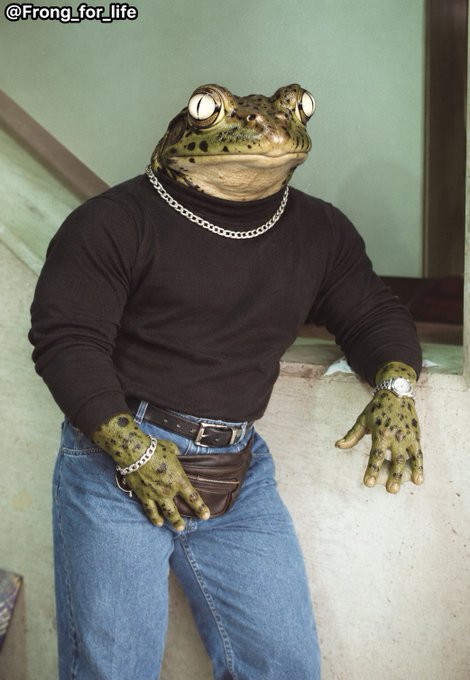 An anthropomorphic frog with a human muscular torso wears a black long-sleeve shirt, jeans, a silver chain necklace, and a wristwatch, leaning casually against a concrete wall.