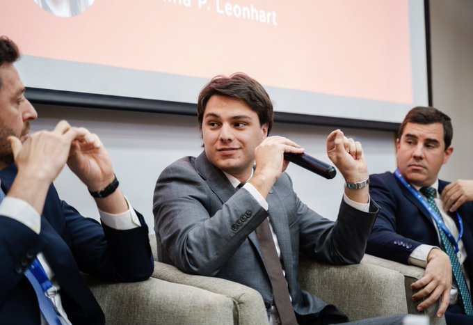 Three men in suits sit on a panel, with the center figure holding a microphone; a projection screen behind them displays the partial name '...P. Leonhart'.