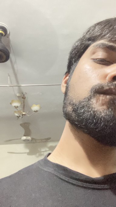 The image features a man with a beard, wearing a black shirt, taking a selfie using his phone's camera. The background of the photo shows a bathroom setting with a sink and a fan hanging from the ceiling. There are also two birds visible in the scene, one on the left side and another closer to the center.