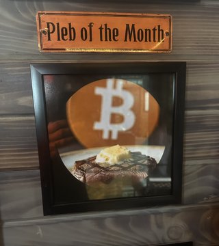 The image features a plate of food with butter, possibly steak or pancakes, placed on top of a wooden table. A sign next to the plate reads "Pleb of the Month", indicating that this month's featured dish is beef buttered pancakes. The background includes a wooden wall and a window, creating an overall rustic atmosphere for the dining experience.