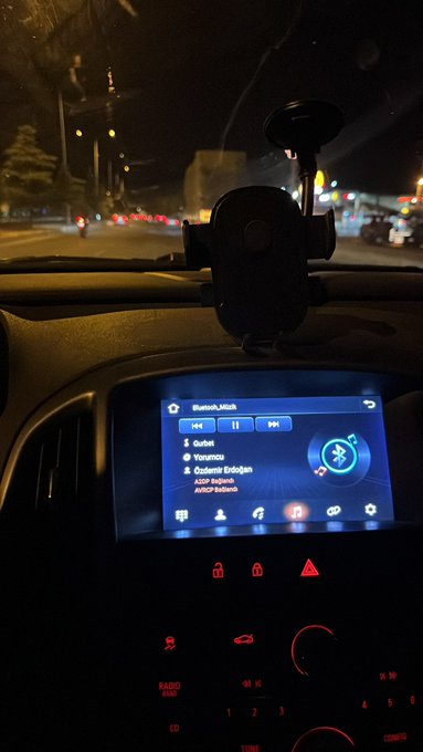 A nighttime photo taken from the passenger seat of a car, showing the dashboard with a Bluetooth audio connection playing "Quitbit" by Yonunca, while blurred street lights are visible through the windshield.