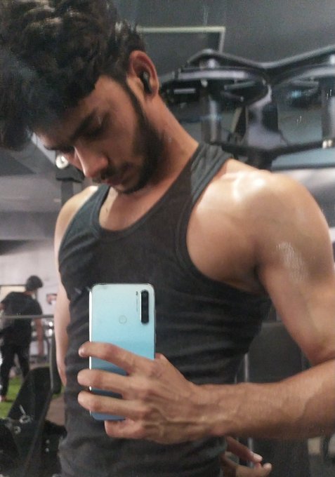 A muscular man in a dark gray tank top takes a mirror selfie with a light blue smartphone in a gym setting.