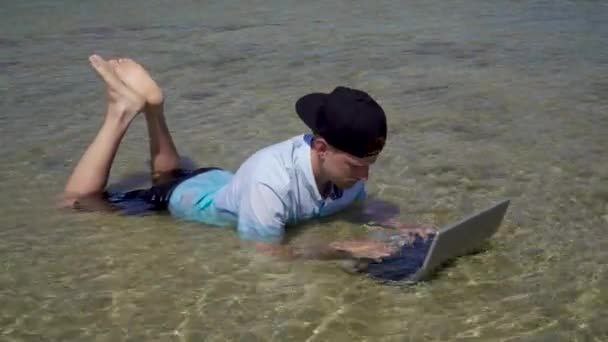 The image shows a man lying face down in shallow water, using his laptop while wearing a hat. He appears to be enjoying himself as he works or relaxes in this unusual setting. The photo also includes text that reads "I'M active and locked in". This phrase suggests that the man is engaged with his work on the laptop despite being in an unconventional environment like the water.