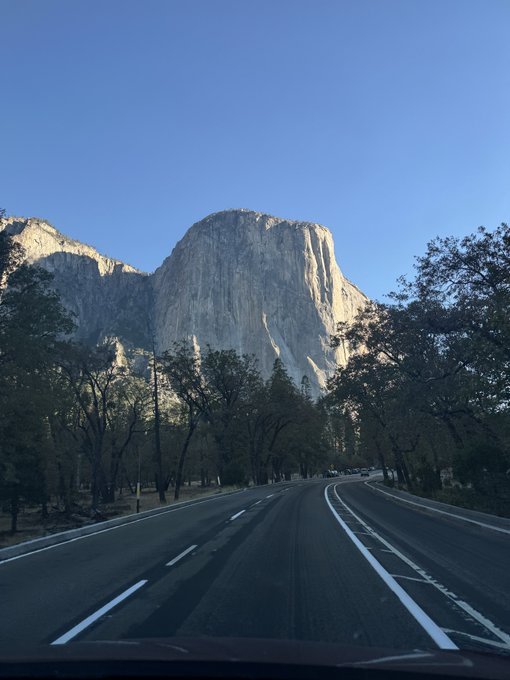 A view from a vehicle on a multi-lane road leading toward a large granite mountain formation under a clear blue sky, likely in Yosemite National Park.