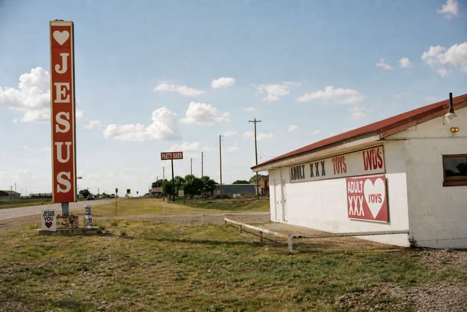 A roadside photograph showing a tall vertical sign reading 'I ♥ JESUS' next to a white building with 'ADULT XXX TOYS' painted in red lettering on its exterior.