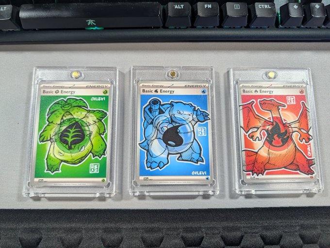 Three Basic Energy Pokémon cards from artist DXLEVI, each featuring a different evolutionary Pokémon (Bulbasaur, Blastoise, and Charizard) in green, blue, and red holographic variants, displayed in protective cases in front of a keyboard.