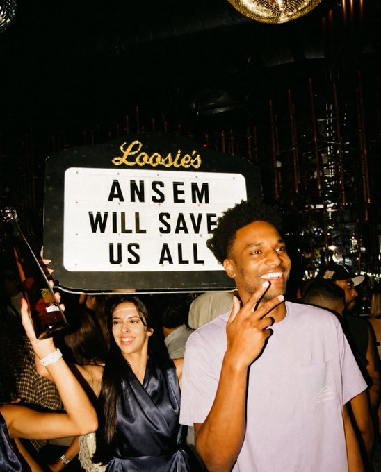 A photograph taken in a dimly lit bar called 'Loosie's', showing a man smiling next to a woman, with a large lit sign behind them that reads 'ANSEM WILL SAVE US ALL'.