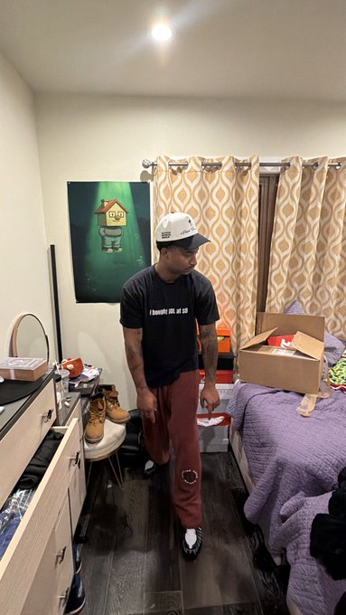 The image shows a man wearing a black t-shirt, red pants, and a white hat standing in a bedroom with a purple bedspread. He has a box of shoes next to him, suggesting he may have recently purchased them or is preparing for an event that requires formal attire. The room also contains other items such as a dresser, mirror, and a chair. In the background, there is a painting on the wall, adding artistic flair to the space.
