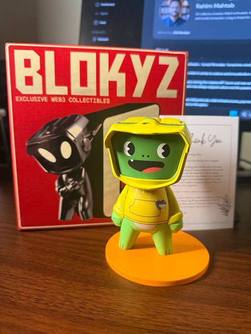 A green Blokyz collectible figure wearing a yellow hoodie stands on an orange base in front of its red "BLOKYZ - Exclusive Web3 Collectibles" packaging box, with a "Thank You" card visible behind it—perfectly capturing the post's sentiment of building a Blokyz collection one piece at a time.