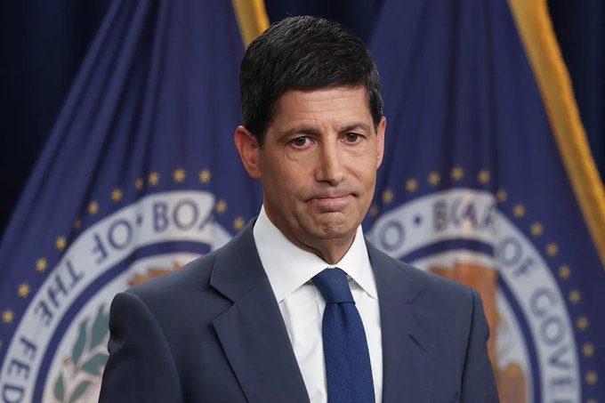 A man in a dark blue suit, white shirt, and blue tie stands at a podium with a serious expression, with partially visible seals of the Board of Governors of the Federal Reserve System behind him. The accompanying post discusses the Federal Reserve's anticipated interest rate hike, and the image of the official corresponds to news related to the Fed's monetary policy decision.
