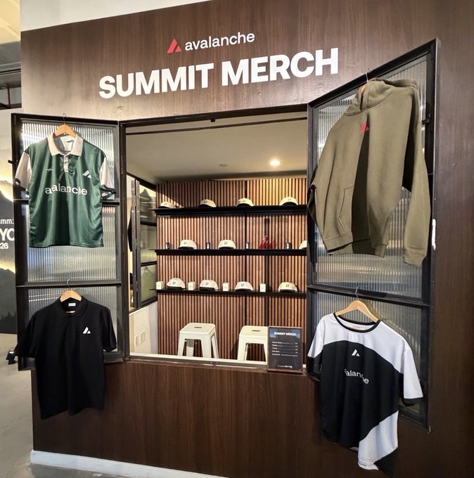 An 'avalanche SUMMIT MERCH' booth displaying branded apparel including a green polo, olive hoodie, black t-shirt, and color-block t-shirt, with hats and accessories on shelves in the center.