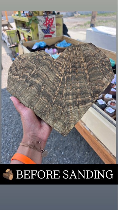 A hand holding a large, rough, heart-shaped piece of wood with prominent wavy grain patterns, with text at the bottom reading 'BEFORE SANDING' next to a wood log emoji.