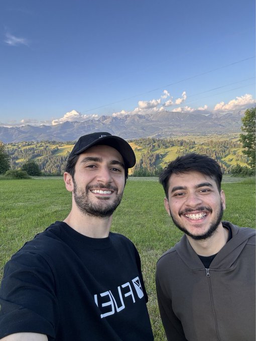 Two men smile in a grassy mountain landscape under a bright blue sky.