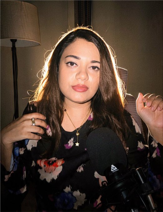 A woman with long brown hair wearing a floral top sits in front of a microphone in a dimly lit room.