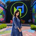 A person in a blue dress and cap stands in front of a colorful graffiti-covered wall featuring a large green music note logo.