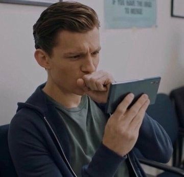 A young man, resembling Tom Holland, sits indoors wearing a dark hoodie, intently focused on a tablet he holds in his hands.