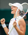 A close-up photograph of a female tennis player wearing a white visor and sleeveless top, shouting in celebration while holding a tennis racket and clenching her fist.