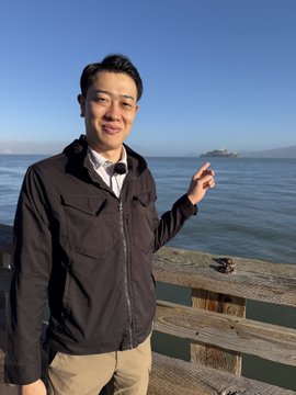 A young Asian man in a black jacket stands on a pier or boardwalk by the water, smiling and pointing with his finger toward something in the distance, with what appears to be Alcatraz Island visible behind him.