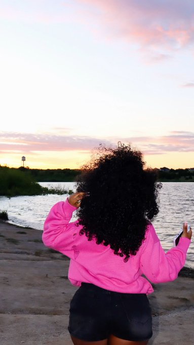 A woman with curly black hair, viewed from behind, wears a bright pink jacket and black shorts while standing on a concrete path overlooking a river at sunset.