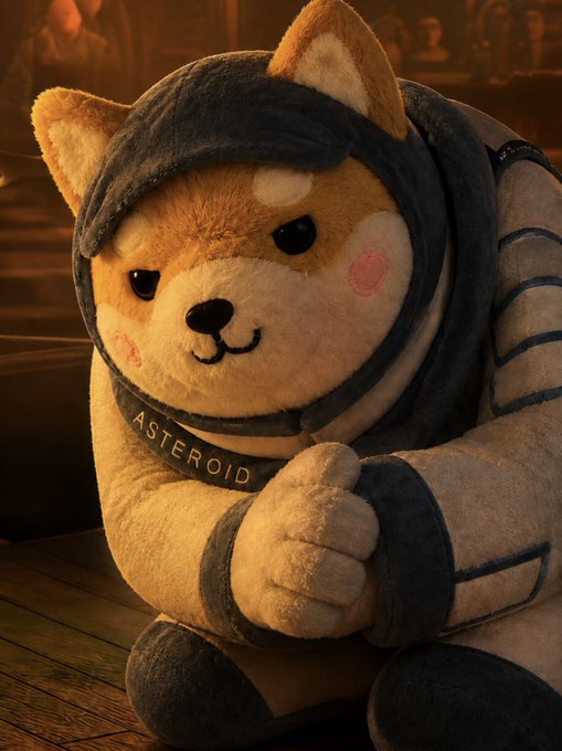 A plush Shiba Inu dog toy wearing a dark hooded outfit and a collar reading "ASTEROID" sits on a wooden surface with a blurred background.
