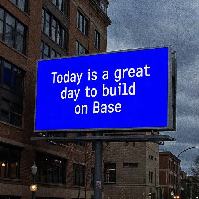 The image features a large blue billboard with white text that reads "Today is a great day to build on Base". The billboard is located near some buildings, suggesting an urban setting. The focus of the image is on the message displayed on the billboard rather than any other objects or elements in the scene.