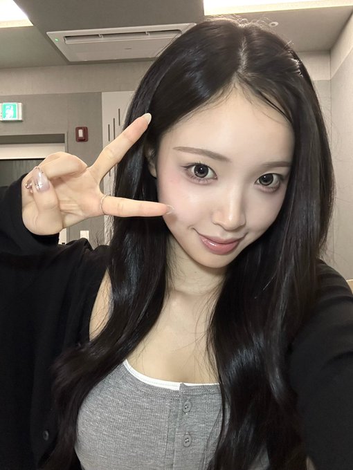 A selfie of a young Asian woman with long dark hair, wearing a black cardigan over a gray top, making a peace sign near her face with a slight smile.