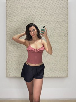 A woman with dark hair takes a mirror selfie with a smartphone while wearing a pink ruffled camisole top and black shorts, standing in front of a textured beige artwork.