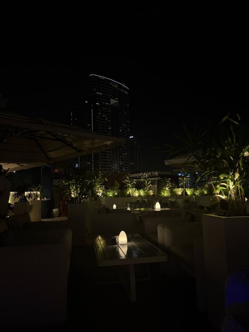 A dimly lit rooftop lounge at night features candles on low tables, lounge seating, and illuminated tropical plants, with a tall skyscraper lit up in the background. The intimate, moody atmosphere complements the post "GN, if you say it back," evoking a quiet, late-night setting perfect for a tender exchange.