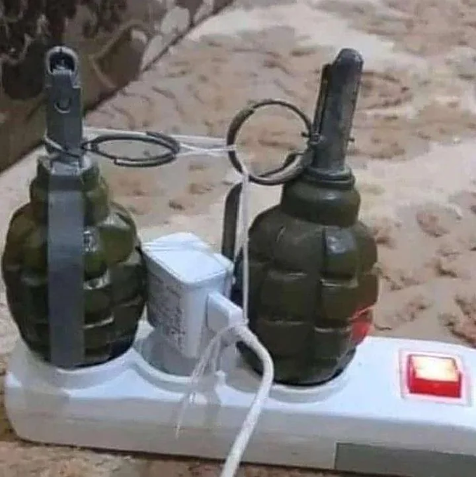 Two green hand grenades rigged with rings and cords, plugged into a white power strip with a red switch, sitting on a surface.