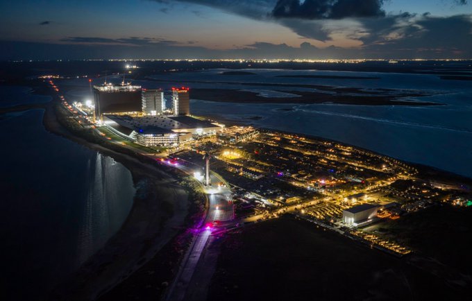 An aerial nighttime view of a brightly lit coastal complex or entertainment venue with a large building, extensive illuminated parking areas, and a bridge, surrounded by water.