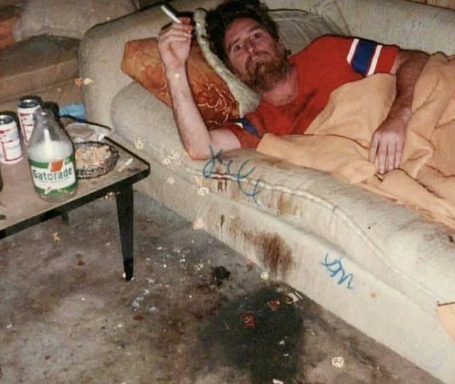 A disheveled man lies on a stained couch holding a cigarette, with a 'Gatorade' bottle and other items on a table nearby.