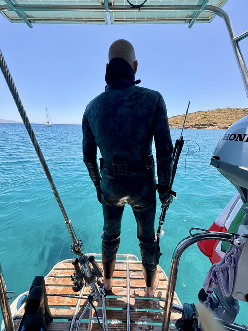 A diver in a wetsuit stands at the back of a boat holding a speargun, ready to plunge into the turquoise water. A Honda outboard motor is visible, and the scene captures a determined spearfishing expedition — a creative alternative livelihood, as the post suggests hoping to bring home a catch.