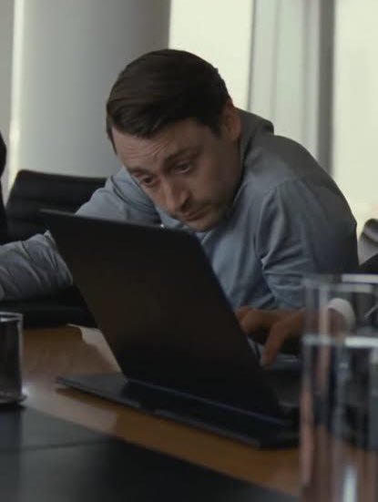 A man in a light blue shirt is intently focused on his laptop screen, leaning forward over a wooden desk with a glass of water and pitcher visible in the foreground.